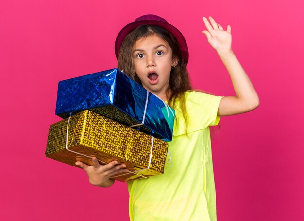 petite fille caucasienne surprise avec un chapeau de fête violet tenant des coffrets cadeaux debout avec une main levée isolée sur un mur rose avec un espace de copie