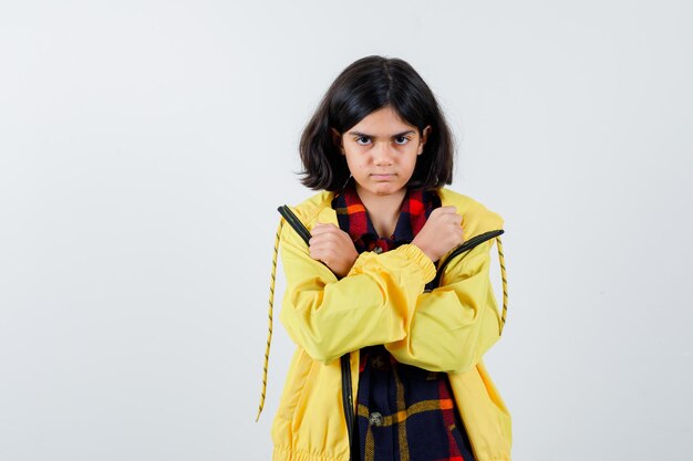 Petite fille en chemise à carreaux, veste montrant les poings croisés sur la poitrine et l'air confiant, vue de face.