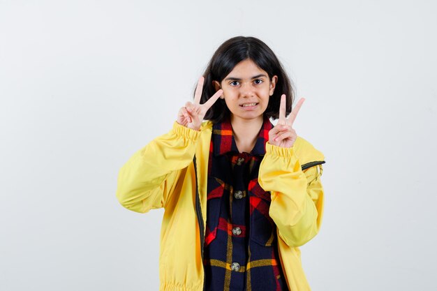 Petite fille en chemise à carreaux, veste montrant le signe de la victoire et semblant chanceuse, vue de face.