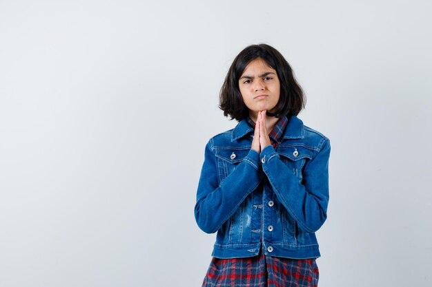 Petite fille en chemise, veste gardant les mains dans un geste de prière et l'air triste, vue de face.