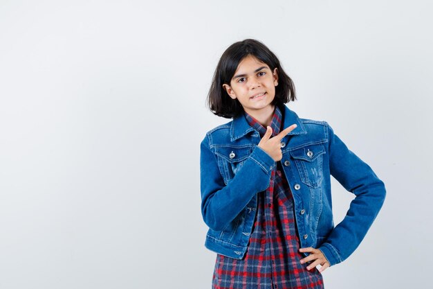 Petite fille en chemise, veste pointant vers le coin supérieur droit et l'air confiant, vue de face.