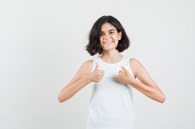 Petite fille en chemisier blanc montrant deux pouces vers le haut et à la vue de face, joyeuse.