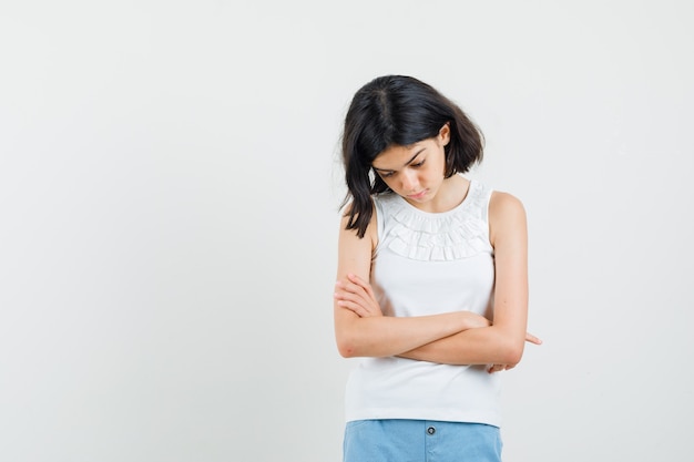 Petite fille en chemisier blanc, short regardant vers le bas avec les bras croisés et regardant pensif, vue de face.