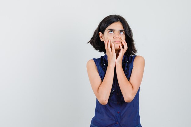 Petite fille en chemisier bleu gardant les mains sur le visage et regardant indécis, vue de face.