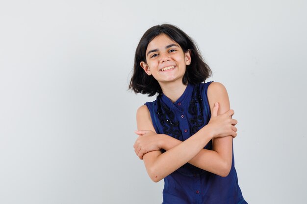 Petite fille en chemisier bleu se serrant dans ses bras et à la joyeuse, vue de face.