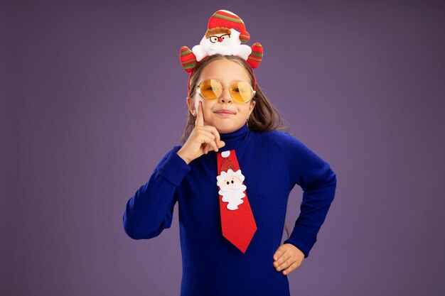 Petite fille en col roulé bleu avec cravate rouge et drôle de jante de Noël sur la tête regardant la caméra avec sourire sur le visage debout sur fond violet