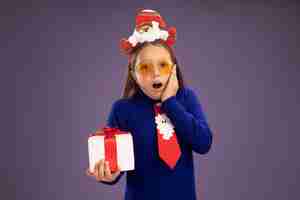 Photo gratuite petite fille en col roulé bleu avec cravate rouge et drôle de jante de noël sur la tête tenant un cadeau regardant la caméra étonné debout sur fond violet