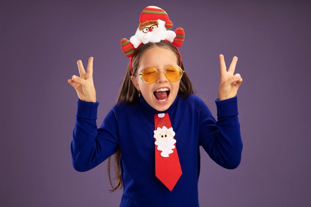 Petite fille en col roulé bleu avec cravate rouge et jante de Noël drôle sur la tête heureux et excité montrant v-sign debout sur fond violet