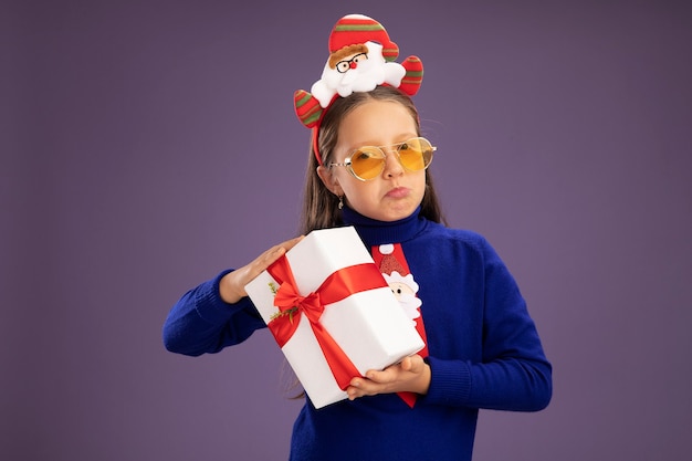 Petite fille en col roulé bleu avec cravate rouge et jante de Noël drôle sur la tête tenant un cadeau en regardant la caméra avec une expression triste en suivant les lèvres debout sur fond violet