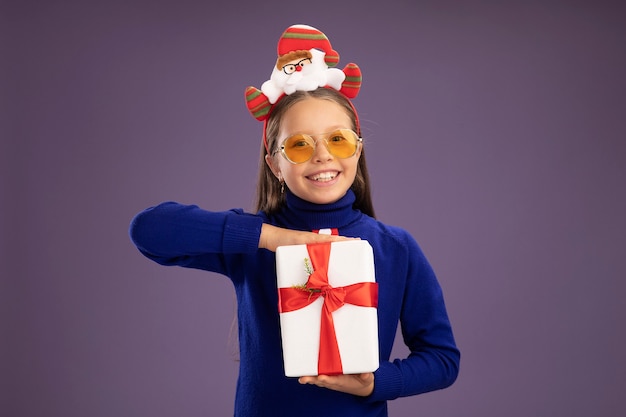 Photo gratuite petite fille en col roulé bleu avec cravate rouge et jante de noël drôle sur la tête tenant un cadeau avec le sourire sur le visage heureux et joyeux debout sur un mur violet