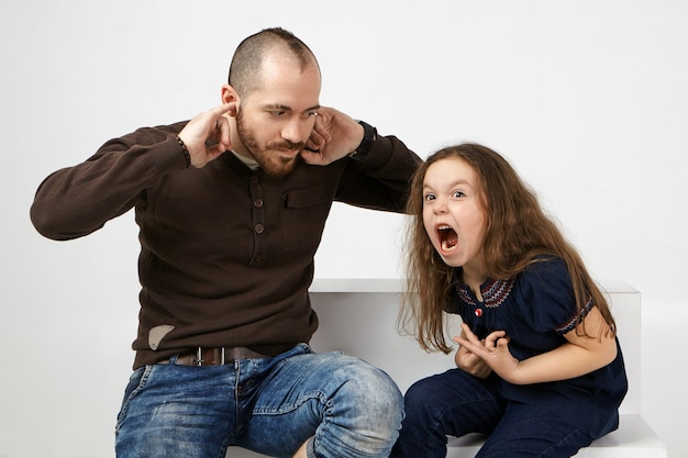 Petite fille en colère avec de longs cheveux lâches criant, se conduisant mal. Frustré, un jeune homme barbu bouchant les oreilles, ne supporte pas les cris ennuyeux de sa fille