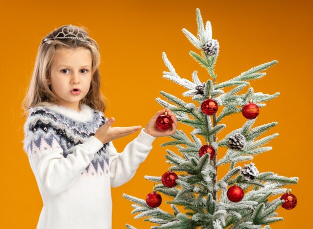 Petite fille confuse debout à proximité de l'arbre de Noël portant diadème avec guirlande sur le cou tenant et points à boule de Noël isolé sur fond orange