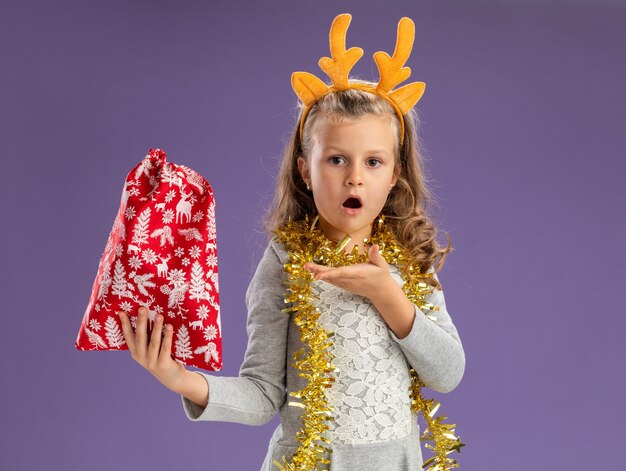 Petite fille confuse portant un cerceau de cheveux de Noël avec guirlande sur le cou tenant et points au sac de Noël isolé sur fond bleu