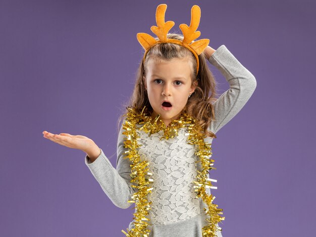 Petite fille confuse portant cerceau de cheveux de Noël avec guirlande sur les points du cou avec la main sur le côté mettant la main derrière la tête isolé sur fond bleu