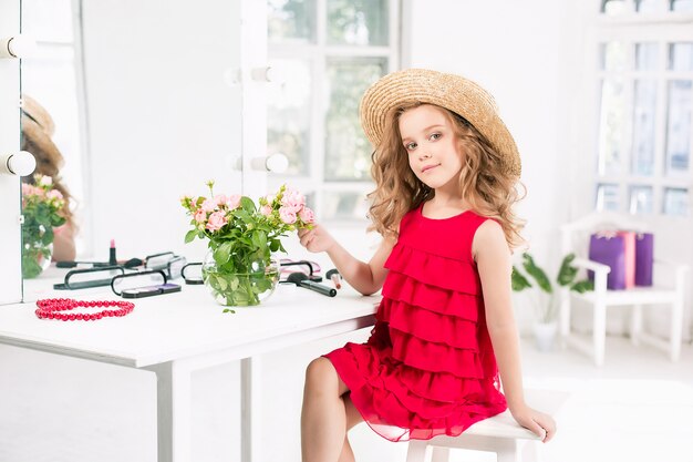 Une petite fille avec des cosmétiques. Elle est dans la chambre de sa mère, assise près du miroir.