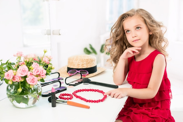 Une petite fille avec des cosmétiques. Elle est dans la chambre de sa mère, assise près du miroir.