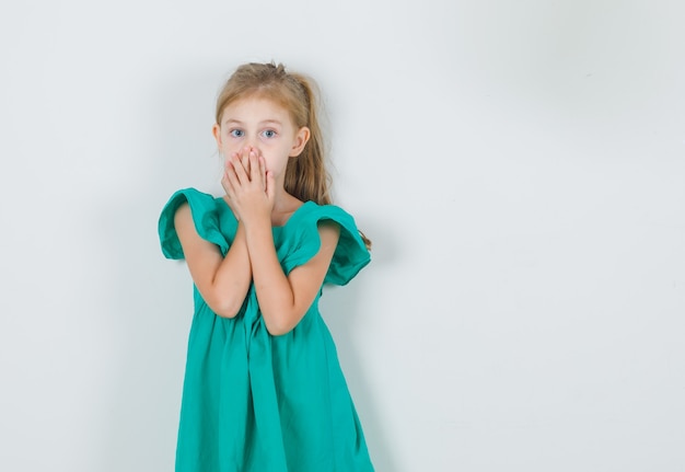 Petite fille couvrant la bouche avec les mains en robe verte et à la surprise. vue de face.