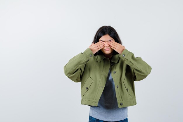 Petite fille couvrant les yeux avec les mains dans un manteau, un t-shirt, un jean et l'air sérieux, vue de face.