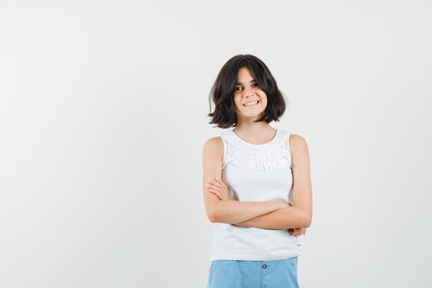 Petite fille debout avec les bras croisés en chemisier blanc, short et à la joyeuse. vue de face.