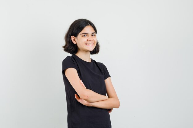 Petite fille debout avec les bras croisés en t-shirt noir et à la joyeuse vue de face.