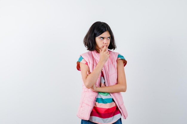 Petite fille debout en pensant pose en t-shirt, gilet bouffant, jeans et l'air pensif. vue de face.