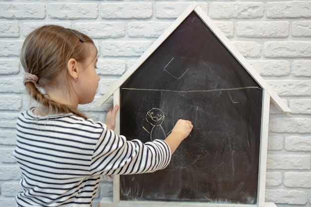Une petite fille dessine un dessin à la craie sur un tableau noir en forme de maison.