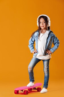 Petite fille élégante dans les écouteurs avec planche à roulettes en vêtements de jeans sur fond orange