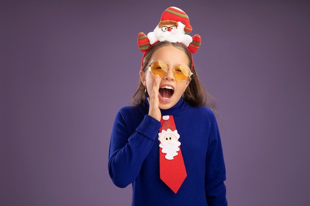 Petite fille émotionnelle en col roulé bleu avec cravate rouge et jante de Noël drôle sur la tête criant avec la main près de la bouche debout sur un mur violet