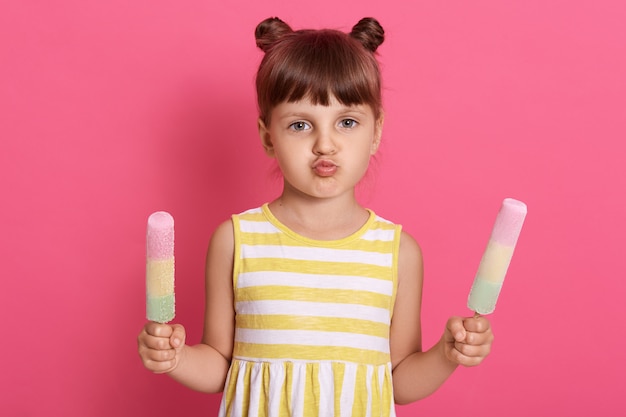 Petite fille enfant tenant des glaces dans les deux mains, avec des lèvres arrondies, enfant de sexe féminin avec deux petits pains isolés sur mur rose.