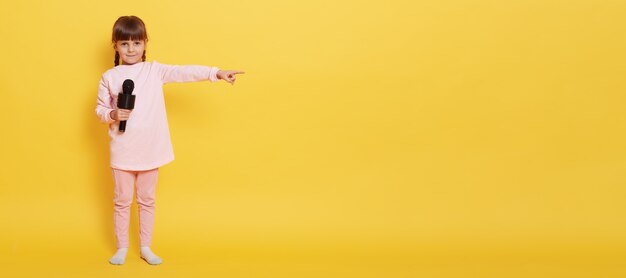 Petite fille européenne avec microphone regarde la caméra tout en tenant le micro, pointe l'index de côté sur un espace vide pour la publicité ou la promotion, charmant chanteur présentant quelque chose au mur jaune.