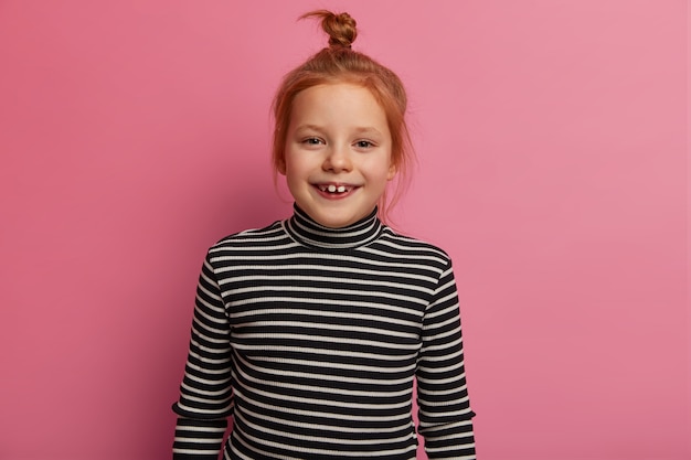 Une petite fille européenne se tient avec désinvolture sur un mur rose pastel, a un chignon de cheveux roux, porte un col roulé à rayures noires et blanches, étant un enfant obéissant, regarde amusé et joyeux, reçoit un beau cadeau