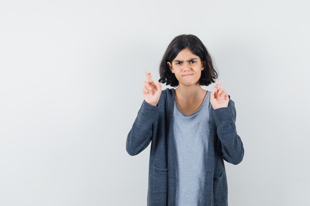 Petite fille en gardant les doigts croisés en t-shirt, veste et à la recherche du doute.