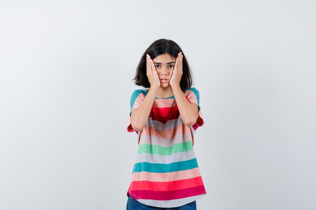 Petite fille gardant les mains sur les joues en t-shirt et ayant l'air impuissante, vue de face.