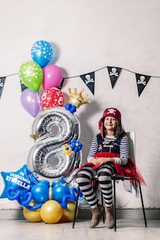 Petite fille habillée en pirate, assise sur une chaise avec des ballons de célébration numéro huit