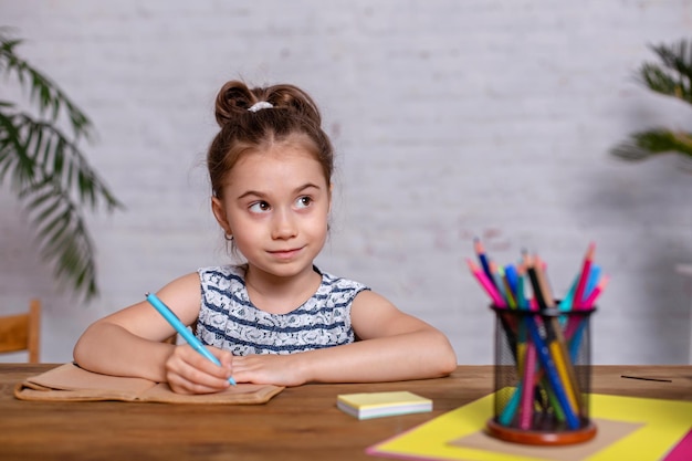 Une petite fille inspirée à la table dessine avec des crayons ou fait ses devoirs. Étudier à l'école primaire. Éducation à domicile