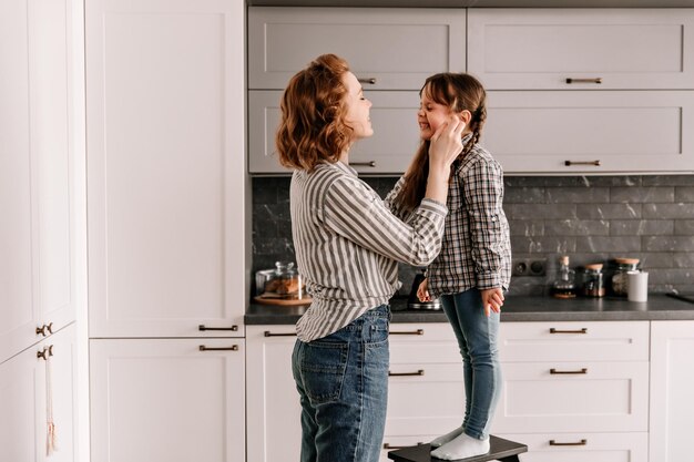 Une petite fille en jeans se tient sur une chaise et rit Maman touche les joues de sa fille