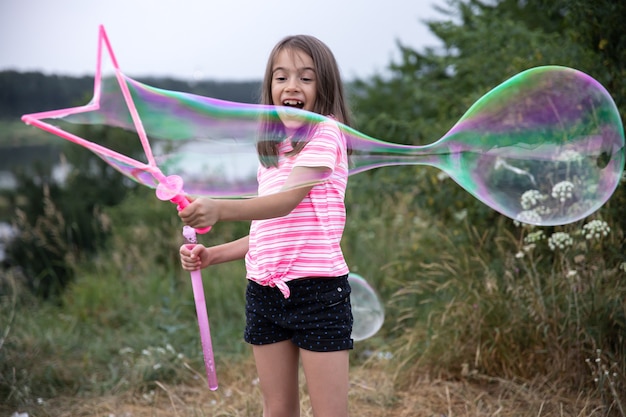 Petite fille joyeuse joue avec de grosses bulles de savon dans la nature.