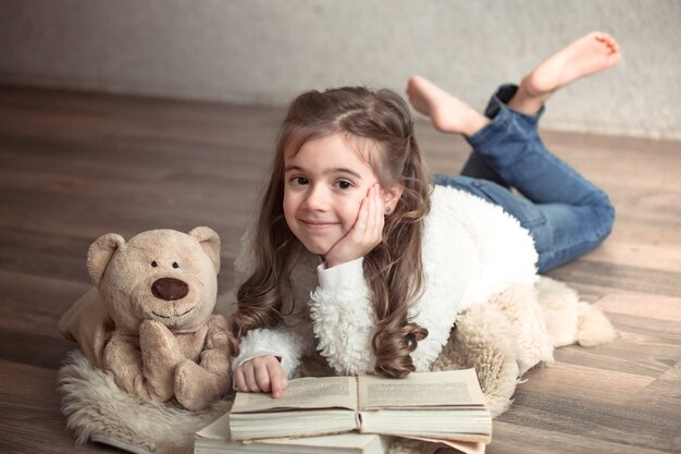 Petite fille lisant un livre avec un ours en peluche sur le sol, concept de détente et d'amitié