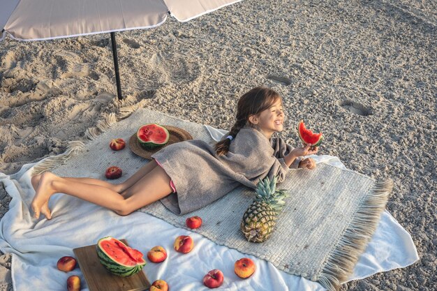 La petite fille mange le fruit se trouvant sur une couverture sur la plage