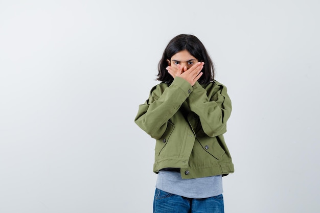 Petite fille en manteau, t-shirt, jeans couvrant la bouche avec les mains et l'air confiant, vue de face.