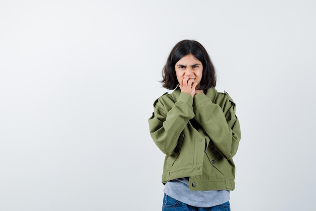 Petite fille en manteau, t-shirt, jeans se rongeant les ongles et l'air sombre, vue de face.