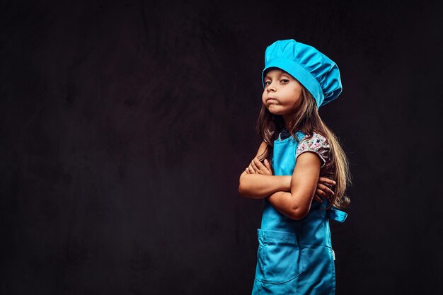 Petite fille mécontente vêtue d'un uniforme de cuisinier bleu debout avec les bras croisés. Isolé sur un fond texturé sombre.