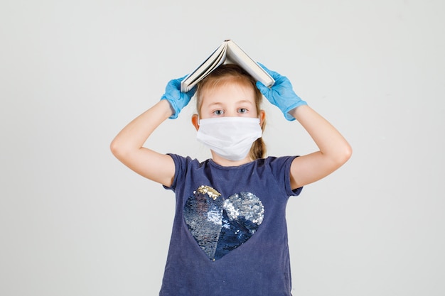 Petite fille mettant un livre sur sa tête en t-shirt, gants et masque vue de face.