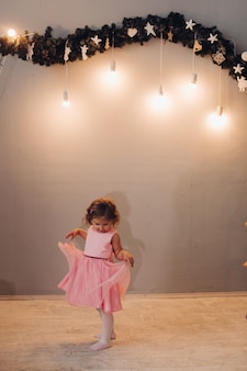 Une petite fille mignonne dans une robe aux cheveux bouclés regarde sa robe près de la guirlande