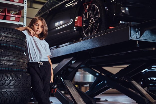 Une petite fille mignonne pose pour un photographe dans un atelier automobile sombre avec une perceuse pneumatique dans les mains.