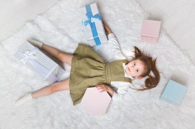 La petite fille mignonne se trouve sur un tapis blanc avec des cadeaux de noël autour.