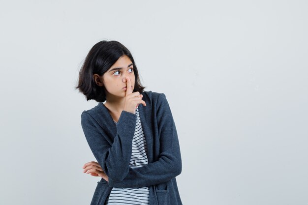 Petite fille montrant le geste de silence en t-shirt, veste et l'air inquiet