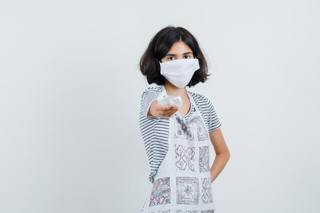 Petite fille offrant un masque médical en t-shirt, tablier