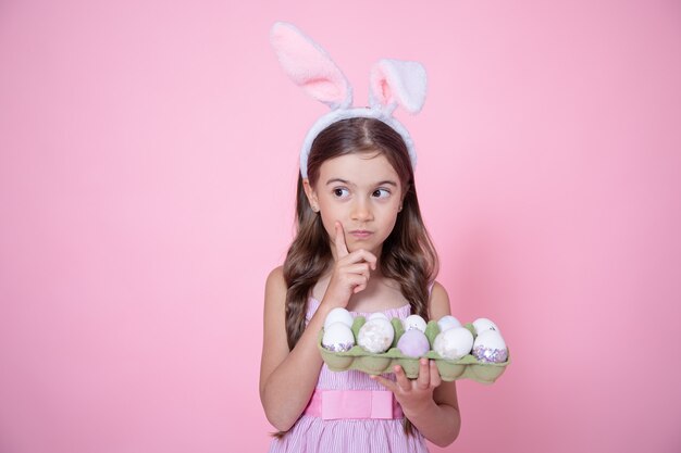 Petite fille avec des oreilles de lapin de Pâques et un plateau d'oeufs dans ses mains sur rose