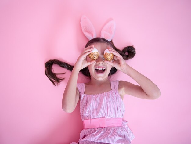 Petite fille avec des oreilles de lapin de Pâques posant avec des oeufs de Pâques festifs allongés sur un mur rose.